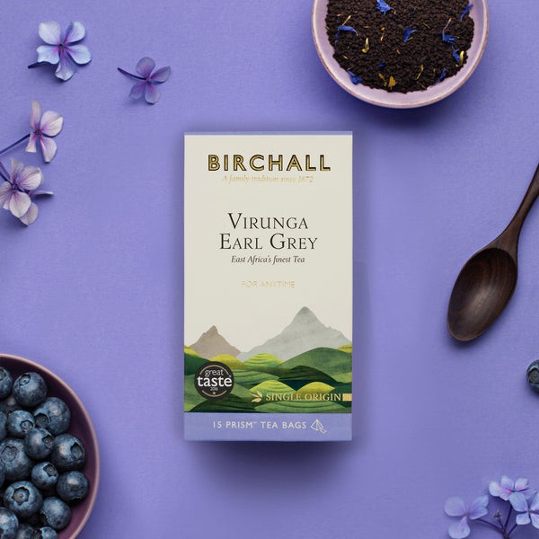 Birds eye view of Birchall Virunga Earl Grey box of 15 prism teabags laid on purple background with tea leaves, spoon, berries and flowers to decorate