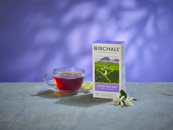 front lifestyle view of Birchall Virunga Earl Grey Box of 15 Prism Teabags. Stood on stone shelf, purple background, glass tea cup with brewed tea in and flowers to decorate